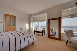 Ein Schlafzimmer mit Bett, Stuhl, Fenster mit Meerblick und Spiegel, der den Raum wiederspiegelt.