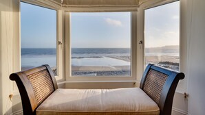 A room with a view of the beach through large windows, a bed with a white mattress, and two chairs with woven backs. - 4 Bed in Amroth (oc-g27016) (Amroth)