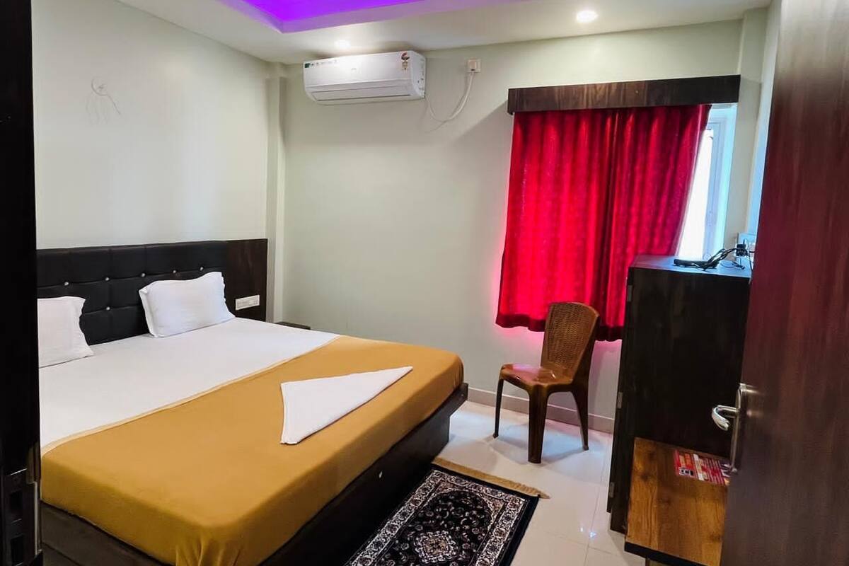 A hotel room with a bed, a desk, a chair, an air conditioner, and a window with red curtains.