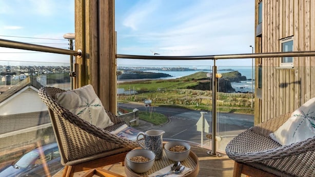 A balcony with wicker furniture, a bowl of cereal, and a mug, overlooking a coastal landscape. - 2 Bed in Newquay (oc-a27276) (Newquay)