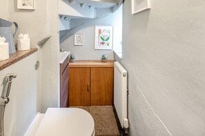 Ein schmales Badezimmer mit einer weißen Toilette, Holzschränken, einem Heizkörper und einer kleinen Ablage mit Pflegeprodukten.