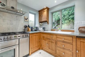 Une cuisine équipée d’une cuisinière à gaz, d’un four, de meubles de rangement en bois, d’une fenêtre donnant sur un espace verdoyant et d’un plan de travail sur lequel sont disposés divers ustensiles de cuisine.