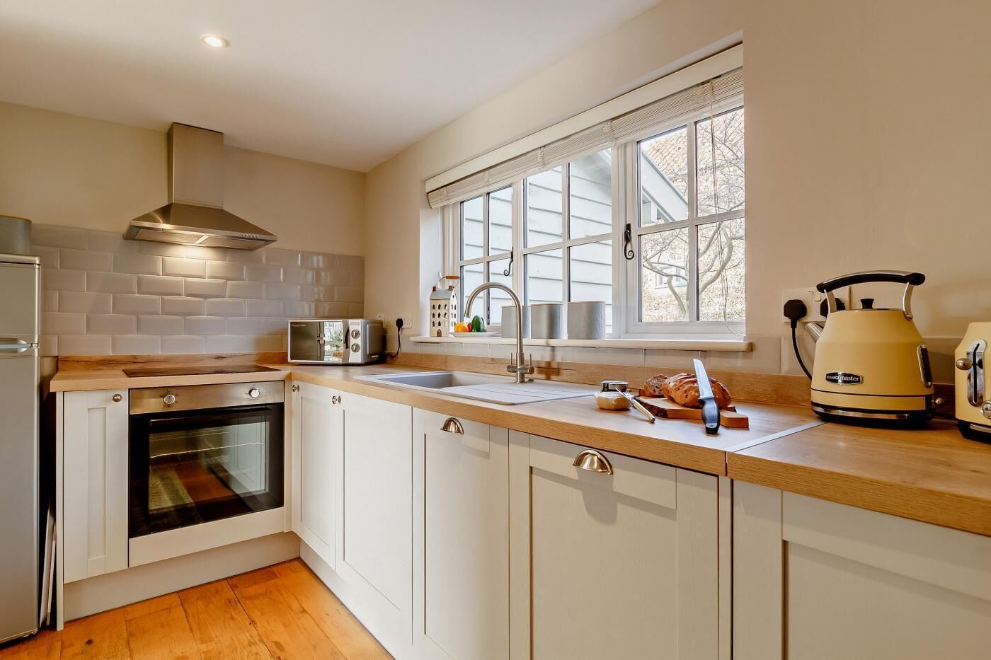 A modern kitchen with white cabinets, a built-in oven, a microwave, a sink, and a window with blinds.