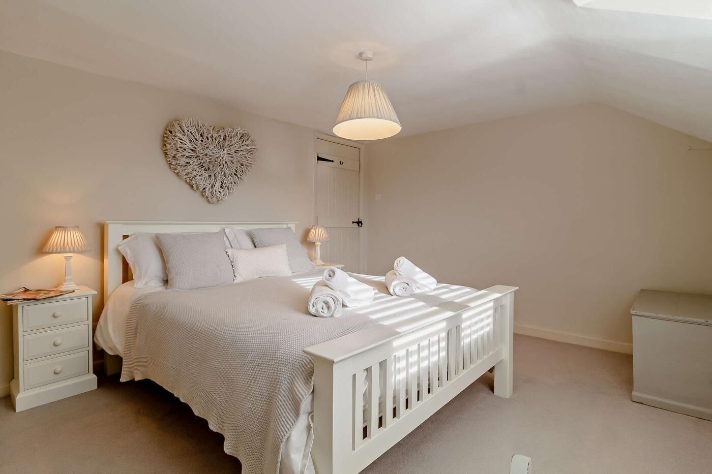 A bedroom with a white bed, bedside tables, a heart-shaped wall decoration, and a ceiling light.