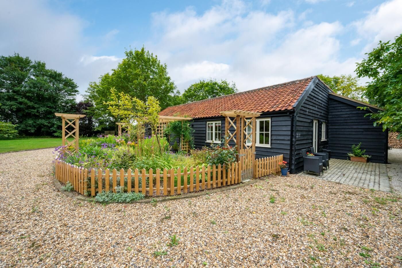 Une maison en bois peinte en noir, avec un toit recouvert de tuiles, entourée d’une allée en gravier et d’une clôture en bois.