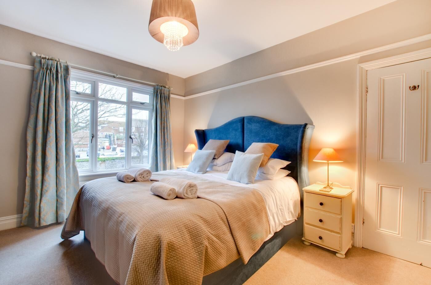 Une chambre à coucher avec un grand lit, une table de chevet, une lampe et une vue sur l’extérieur par une fenêtre.