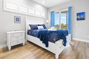 Une chambre à coucher avec un lit blanc, une table de chevet, une fenêtre avec des rideaux bleus et une vue sur l’extérieur.