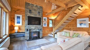 L’intérieur d’un chalet accueillant, avec une cheminée en pierre, un téléviseur à écran plat, un escalier et un canapé en cuir blanc.