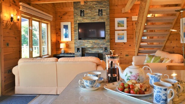 L’intérieur chaleureux d’un chalet avec un canapé, une table basse sur laquelle sont posées des tasses à thé et une assiette de fraises, une cheminée et un téléviseur fixé sur un mur en pierre.