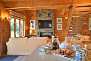 L’intérieur chaleureux d’un chalet avec un canapé, une table basse sur laquelle sont posées des tasses à thé et une assiette de fraises, une cheminée et un téléviseur fixé sur un mur en pierre.
