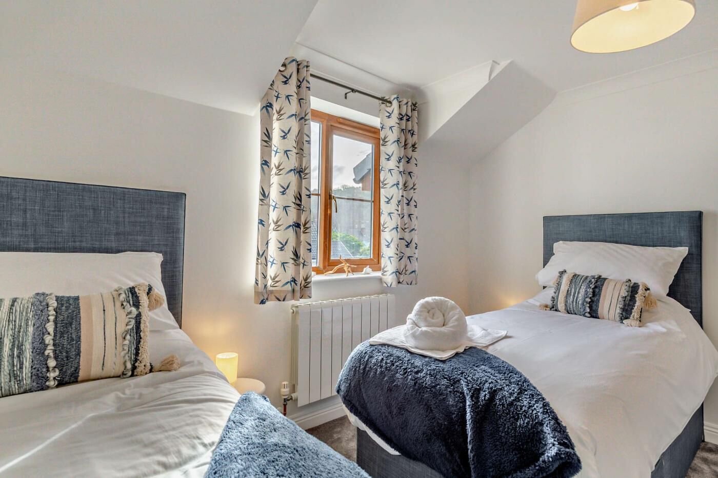 A bedroom with two beds, a window with curtains, a radiator, and a ceiling light.