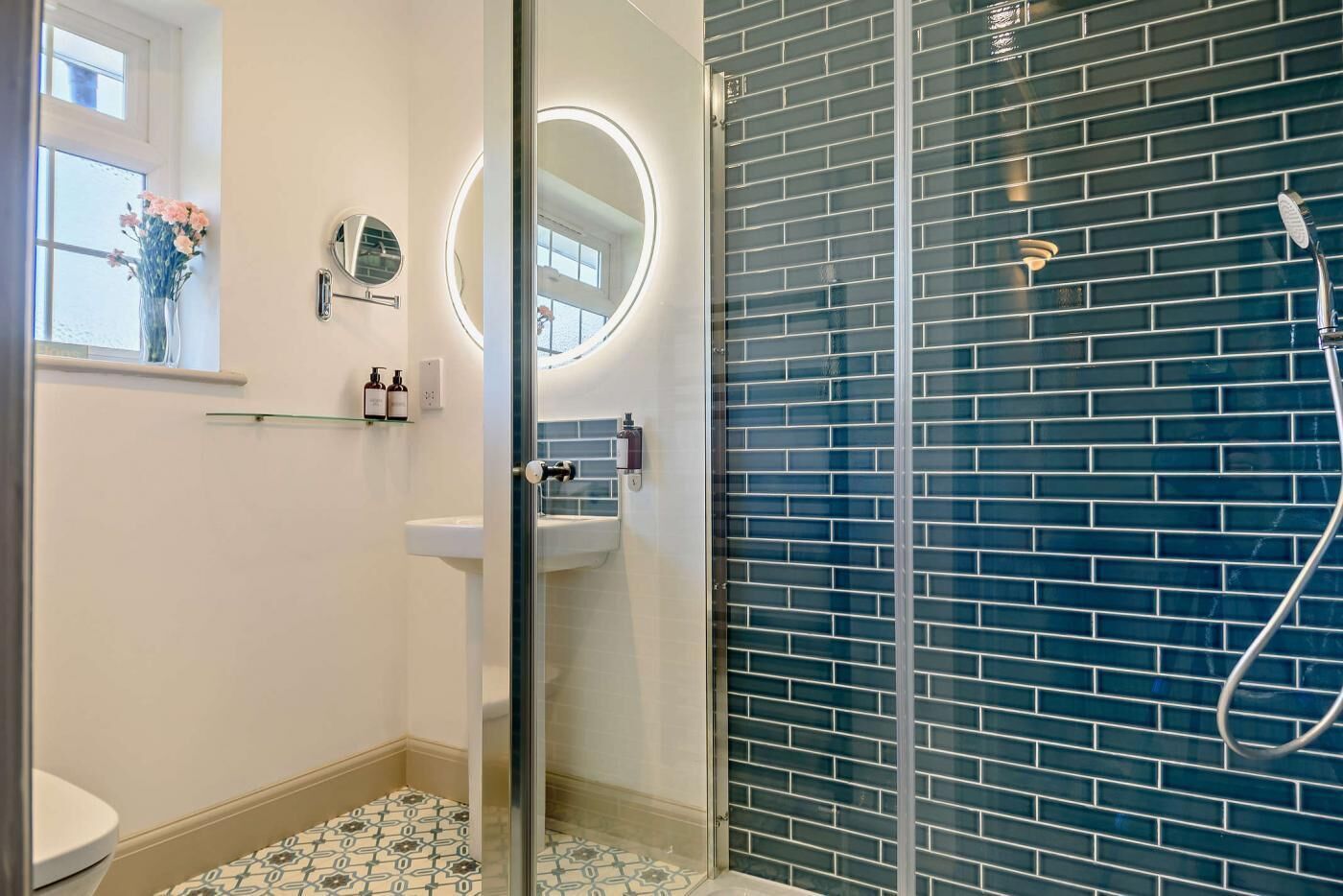 A bathroom with a glass shower enclosure, a sink, and a mirror with a round light.