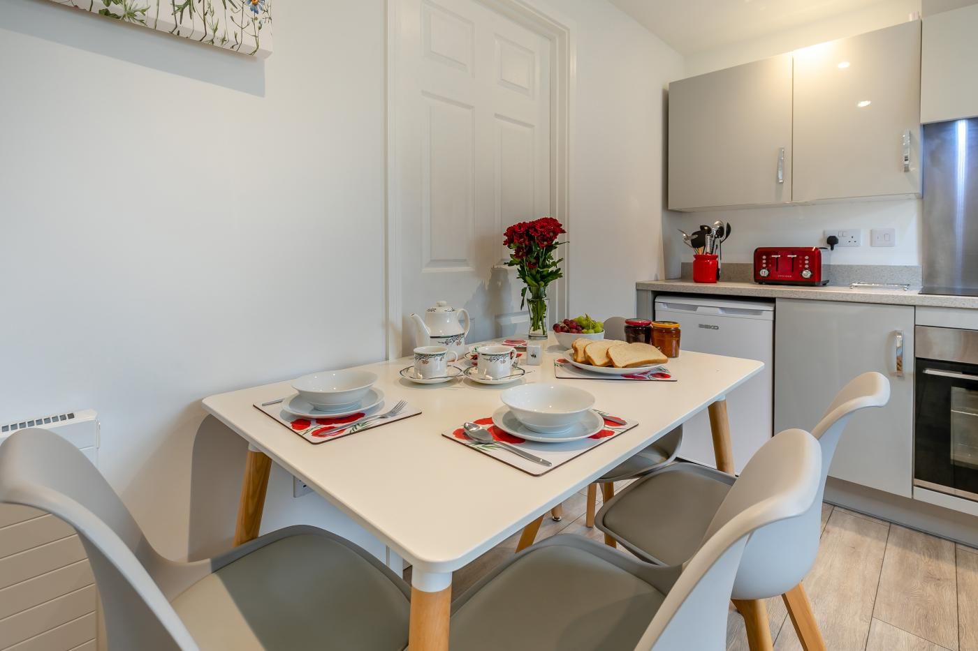A modern kitchen with white cabinets, a stainless steel refrigerator, and a dining table set for two with plates, cups, and a vase of flowers.
