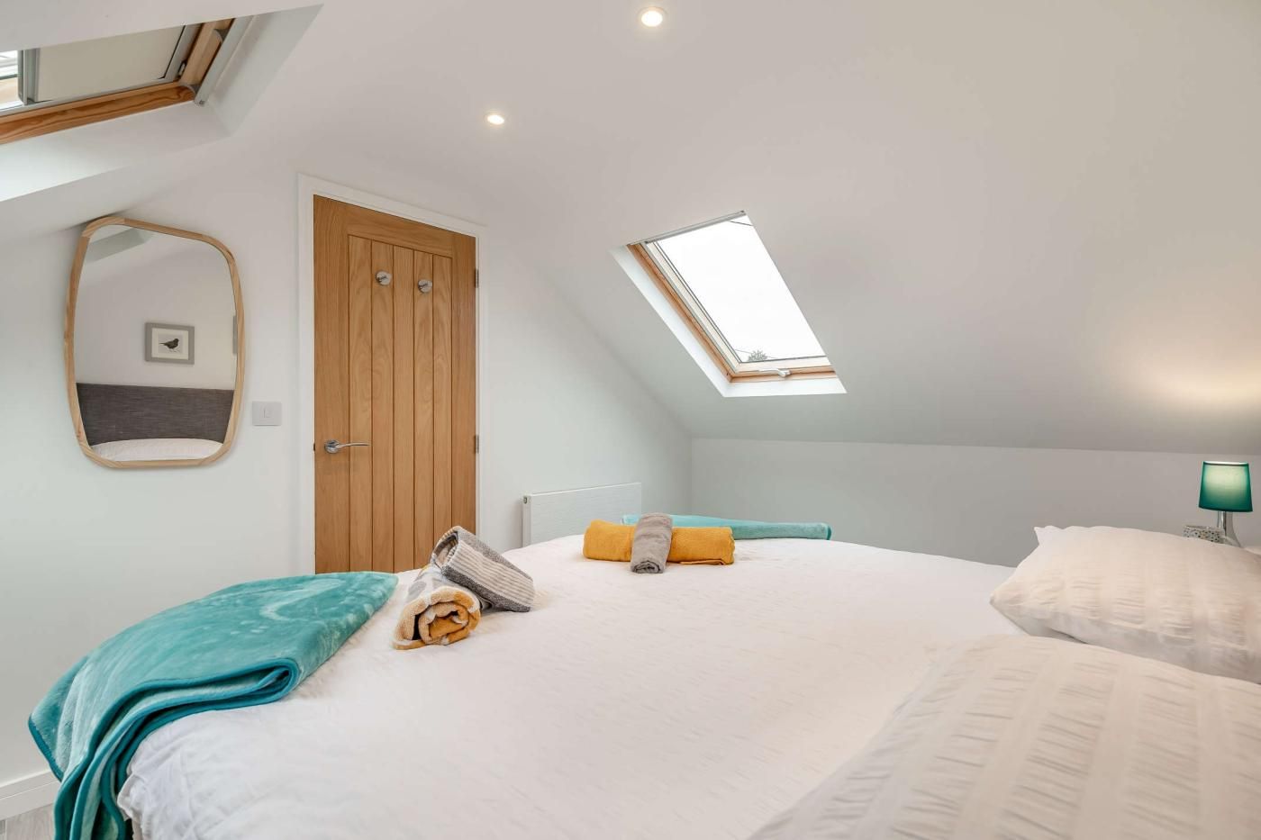 A bedroom with a skylight, a bed with pillows, a bedside table with a lamp, and a wooden door.