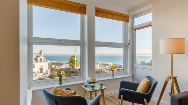 Ett rum med två stora fönster som erbjuder utsikt över havet och byggnaderna. Det finns två blå fåtöljer med gula kuddar, ett litet bord med en vas med blommor och en golvlampa.