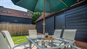 Une table avec un plateau en verre, quatre chaises, un parasol vert et un assortiment de mets pour le petit-déjeuner comprenant des œufs, du bacon et des fruits.