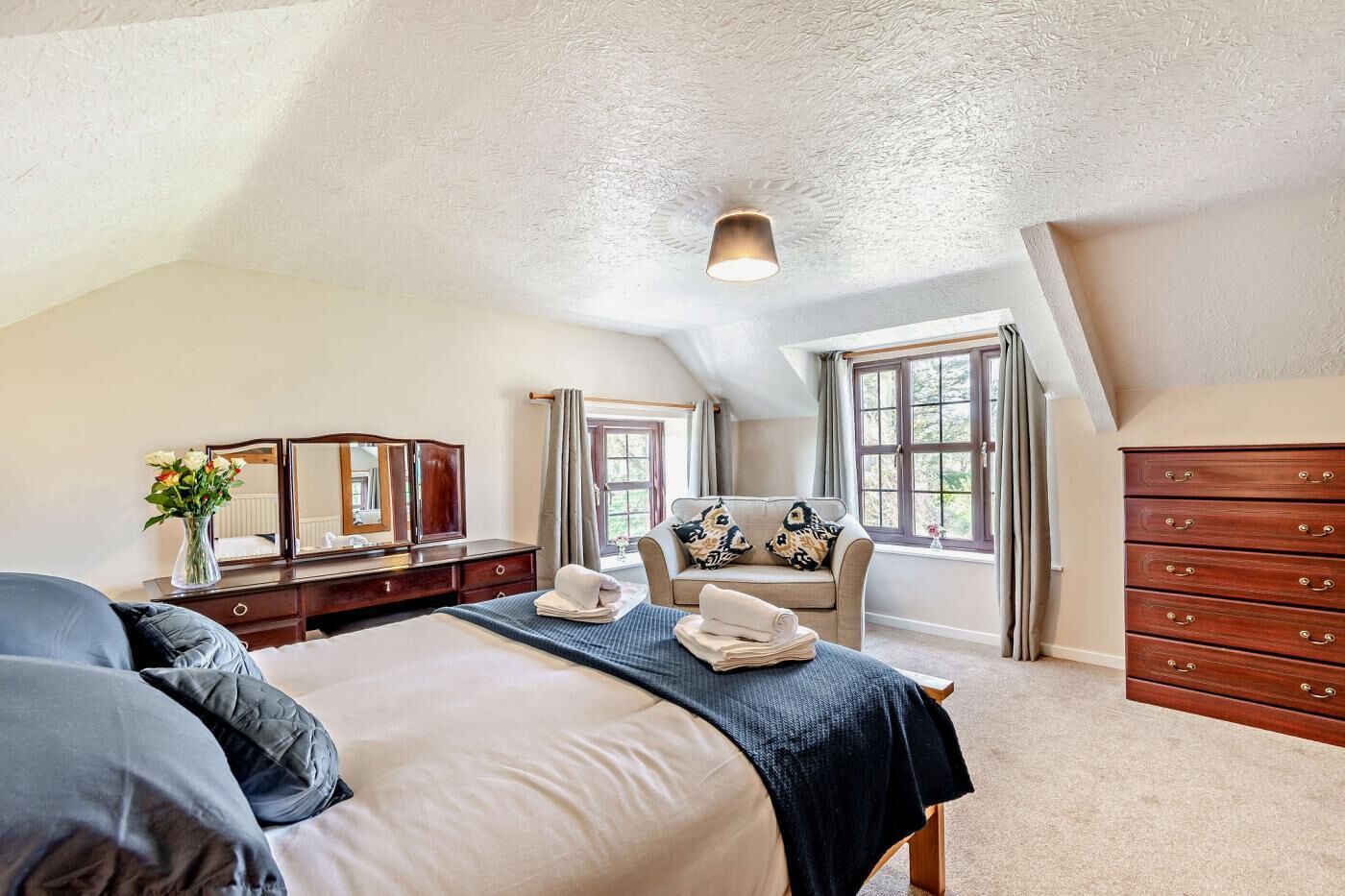 A bedroom with a large bed, a dresser, a vanity with a mirror, a chair, and a window with curtains.