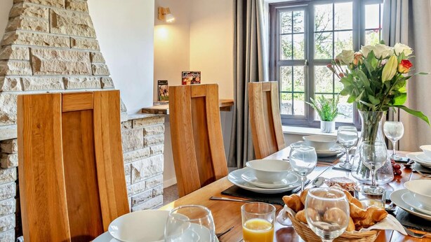 Un comedor con pared de piedra, sillas de madera, una mesa con platos, vasos y un florero.