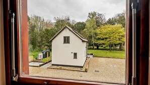 Uitzicht door een raam op een klein wit gebouw met een puntig dak, omringd door bomen en een grindpad.
