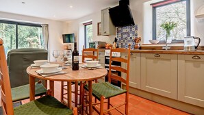 Een eethoek met een ronde tafel, stoelen en een wijnfles. Er is een keuken met een fornuis, spoelbak en keukenkastjes.