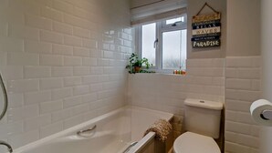 A bathroom with white tiled walls, a bathtub, a toilet, and a small window with a view. - 2 Bed in Southwold (oc-p27380) (Southwold)