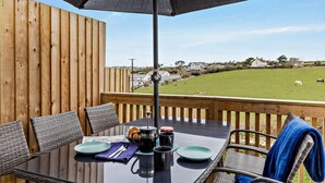 Eine Holzterrasse mit einem für zwei Personen gedeckten Tisch, einem schwarzen Sonnenschirm und Blick auf ein grünes Feld mit Pferden.