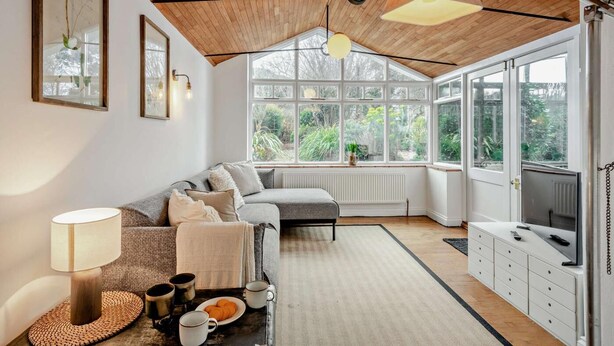 Ein gemütliches Wohnzimmer mit Holzbalkendecke, einer Couch, einem Couchtisch mit Tassen und Gebäck sowie einem großen Fenster mit Blick auf Grünflächen.