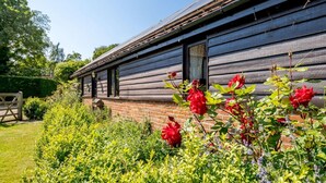 Een rustiek houten gebouw met een bakstenen fundering, omgeven door weelderige begroeiing en bloeiende planten.