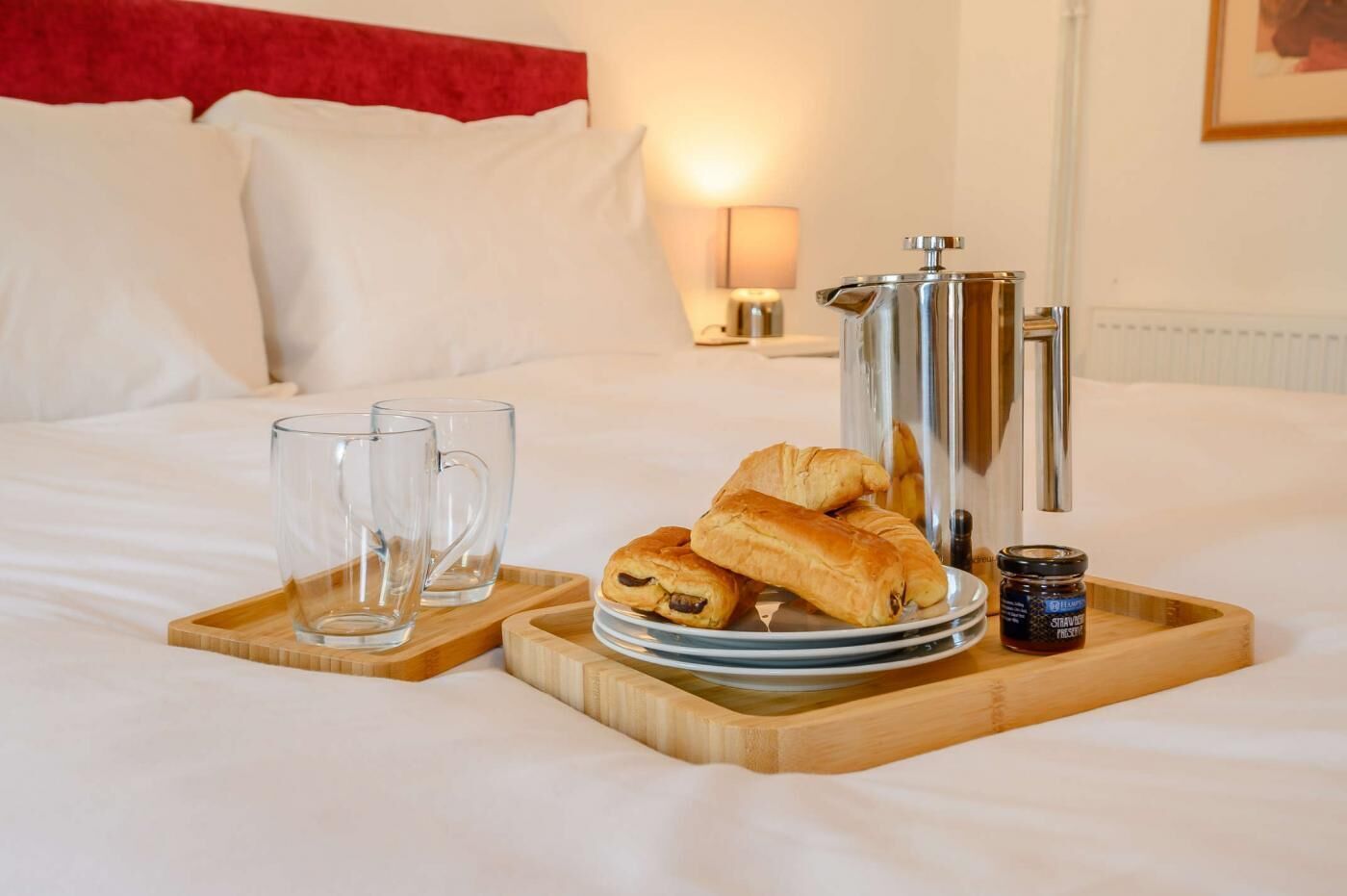 A neatly made bed with a tray of croissants, a jar of jam, and two glasses of water.