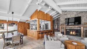 A spacious living area with a wooden dining table, a stone fireplace, and a large TV mounted on the wall. - Ski-In/Out Lodge | Stratton Mountain (Stratton)