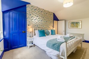 A bedroom with a white bed, teal pillows, a blue door, and a floral wallpaper. - 3 Bed in Kelling (oc-w27424) (Kelling)
