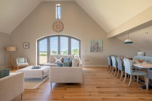 Un loft espacioso con techos altos, una zona de comedor con una mesa larga y sillas, un rincón de estar acogedor con un sofá y sillones, y un amplio ventanal de arco que ofrece vistas al campo.