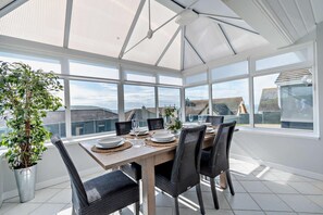 A sunlit dining area with a table set for four, large windows, and a potted plant. - 2 Bed in Gwbert (oc-t27375) (Gwbert)