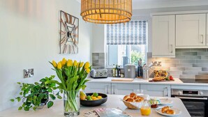 Een moderne keuken met witte kasten, een groot eiland met ontbijtartikelen, een rieten hanglamp en een raam met jaloezieën.