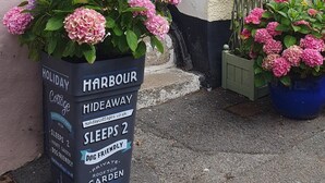 A potted plant with pink flowers and a sign indicating a holiday cottage named "Harbour Hideaway" with amenities like sleeping 2, dog-friendly, and a private garden. - 1 Bed in Mevagissey (oc-h27460) (Mevagissey)