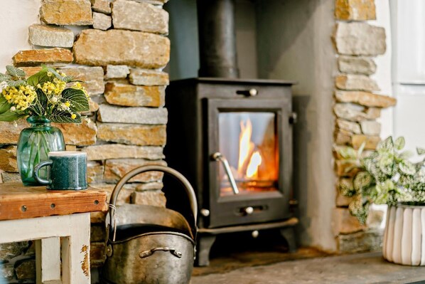 A stone fireplace with a wood-burning stove, a green mug, a metal kettle, and a vase with flowers.