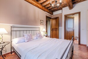 A bedroom with a bed, bedside tables, a wooden ceiling, and a door leading to the bathroom. - Short Rentals Siena-Le Rose, apartment with pool nestled in the Crete Senesi (Toscana)