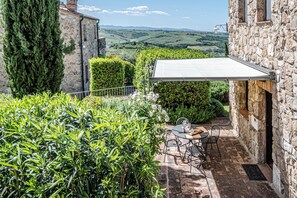 A stone building with a patio area featuring a table and chairs, surrounded by lush greenery and a scenic view of rolling hills. - Short Rentals Siena-Le Rose, apartment with pool nestled in the Crete Senesi (Toscana)
