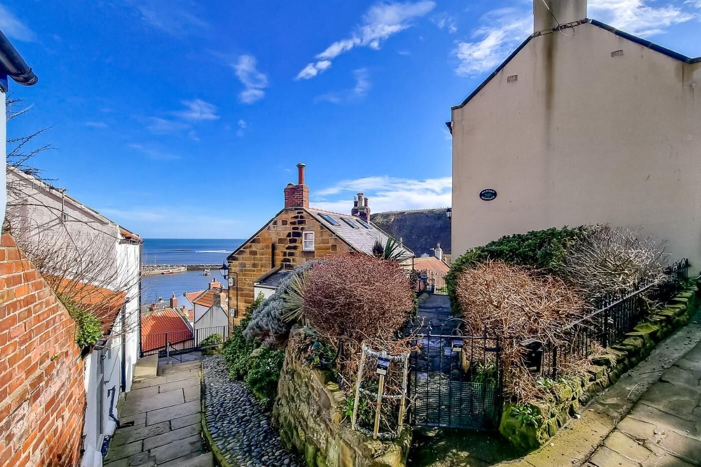 A narrow street with stone walls, a stone building with a red roof, and a view of the sea.