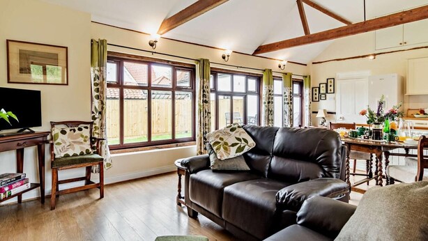 A living room with a leather sofa, wooden coffee table, and a television on a wooden stand. There are large windows with curtains, a kitchen area in the background, and a dining table with chairs.
