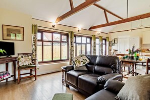 A living room with a leather sofa, wooden coffee table, and a television on a wooden stand. There are large windows with curtains, a kitchen area in the background, and a dining table with chairs.