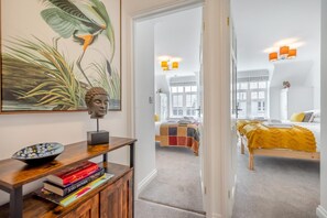 Une chambre d’hôtel avec une grande fenêtre, un lit recouvert d’une couverture aux couleurs vives, et une table de chevet avec des livres et un bol décoratif.