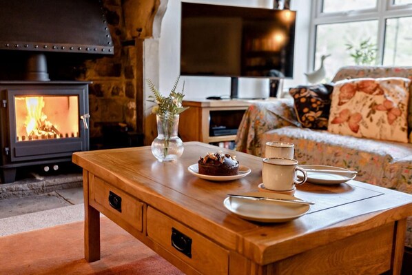Ein gemütliches Wohnzimmer mit Kaminofen, einem Holztisch mit Speisen und Tassen sowie einem blumigen Sofa.