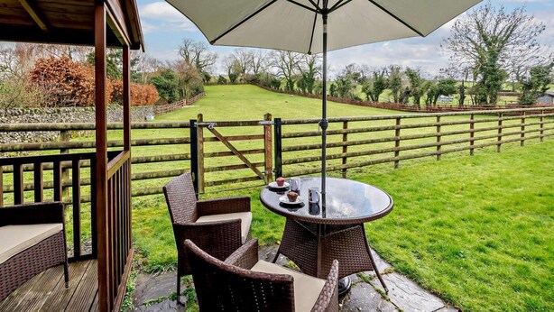 Une terrasse extérieure aménagée avec des meubles en osier et une table placée sous un grand parasol, donnant sur une pelouse délimitée par une clôture en bois.
