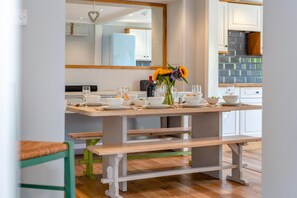 A dining area with a wooden table set for four, a vase with flowers, and a kitchen in the background. - 4 Bed in St. Levan (oc-b27515) (Saint Levan)