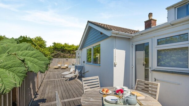 Eine Holzterrasse mit einem runden Esstisch, ein kleines Gebäude mit blauer Fassade und eine große, grüne Pflanze.