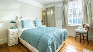 Une chambre à coucher avec un lit, une table de chevet, une chaise, une fenêtre avec des rideaux et un radiateur.