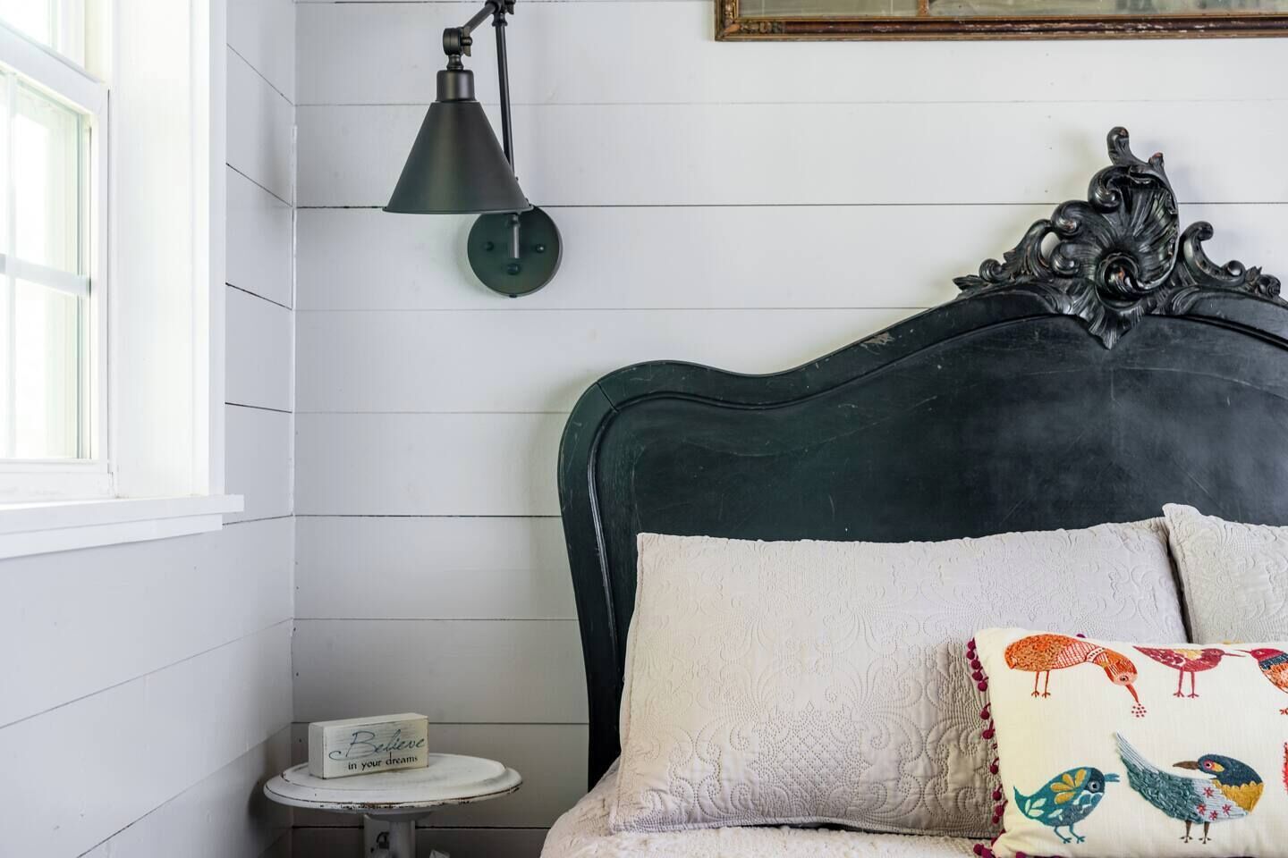 A bedroom with a dark green headboard, a white wall, a small bedside table with a lamp, and a decorative pillow with bird patterns.