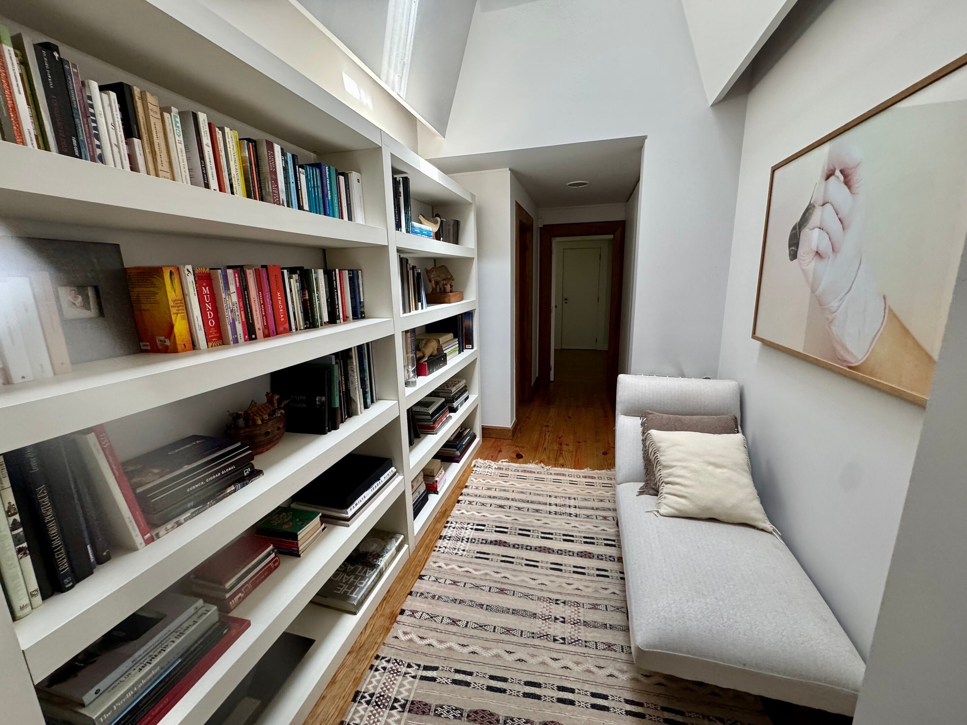 Una habitación con una estantería blanca llena de libros, una alfombra con diseño y un sofá gris con un cojín.