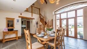 A dining area with a wooden table set for four, surrounded by chairs, a staircase, and a large window with a view. - 3 Bed in Okehampton (oc-h27530) (South Tawton)